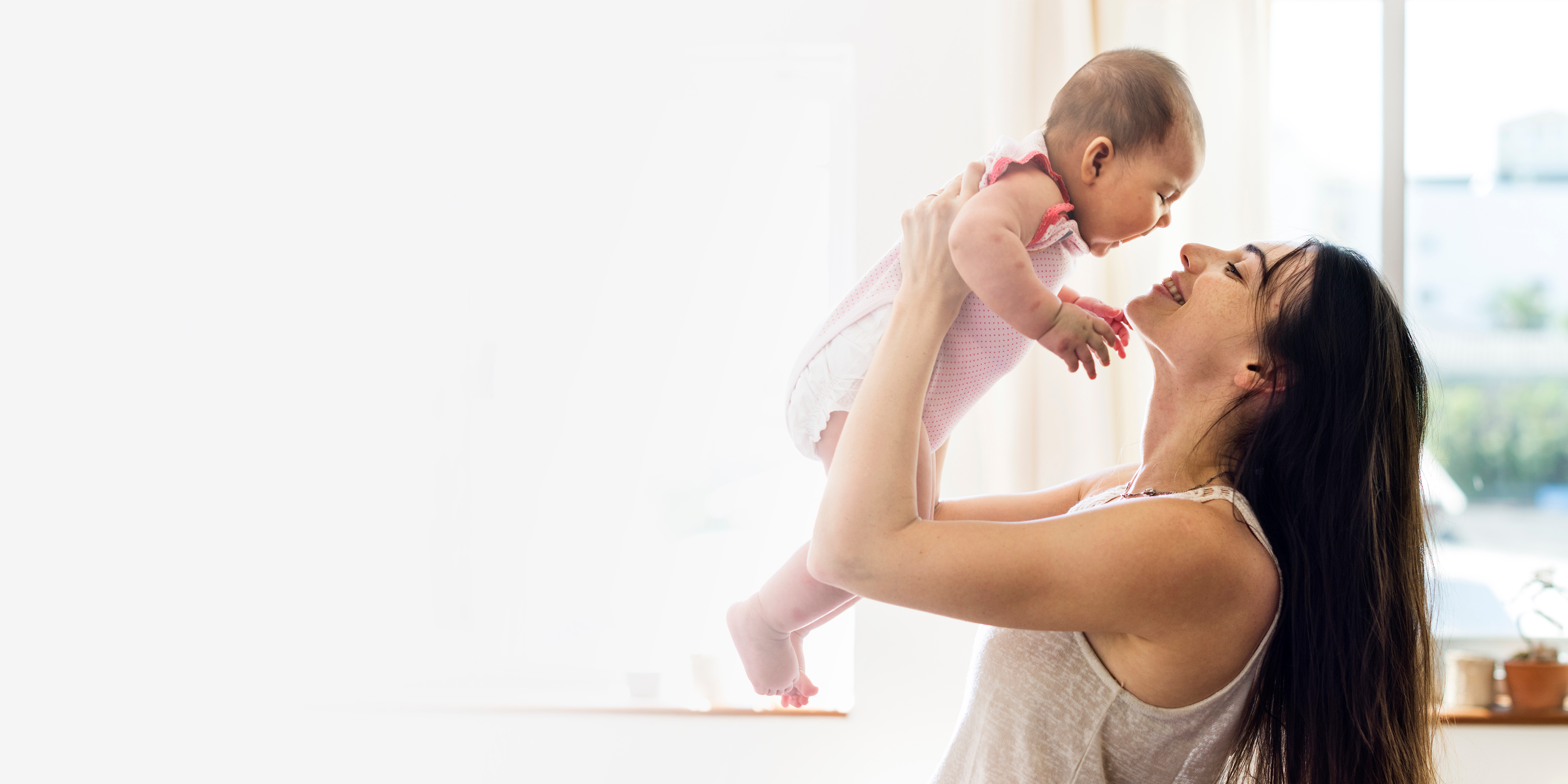 A mother joyfully lifting her baby overhead in a sunlit room